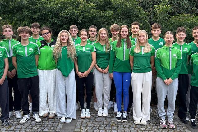 Gruppenbild von 16 jungen Sportlern in gr&uuml;nen Trikots, stehend vor einer gr&uuml;nen Hecke auf einem gepflasterten Weg.