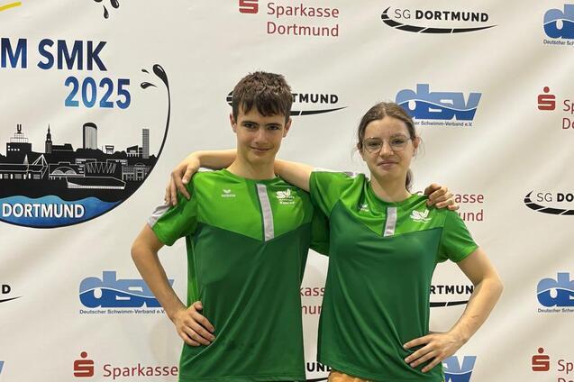 Zwei junge Schwimmer in gr&uuml;nen T-Shirts posieren vor einem Banner der Deutschen Meisterschaften 2025 in Dortmund.
