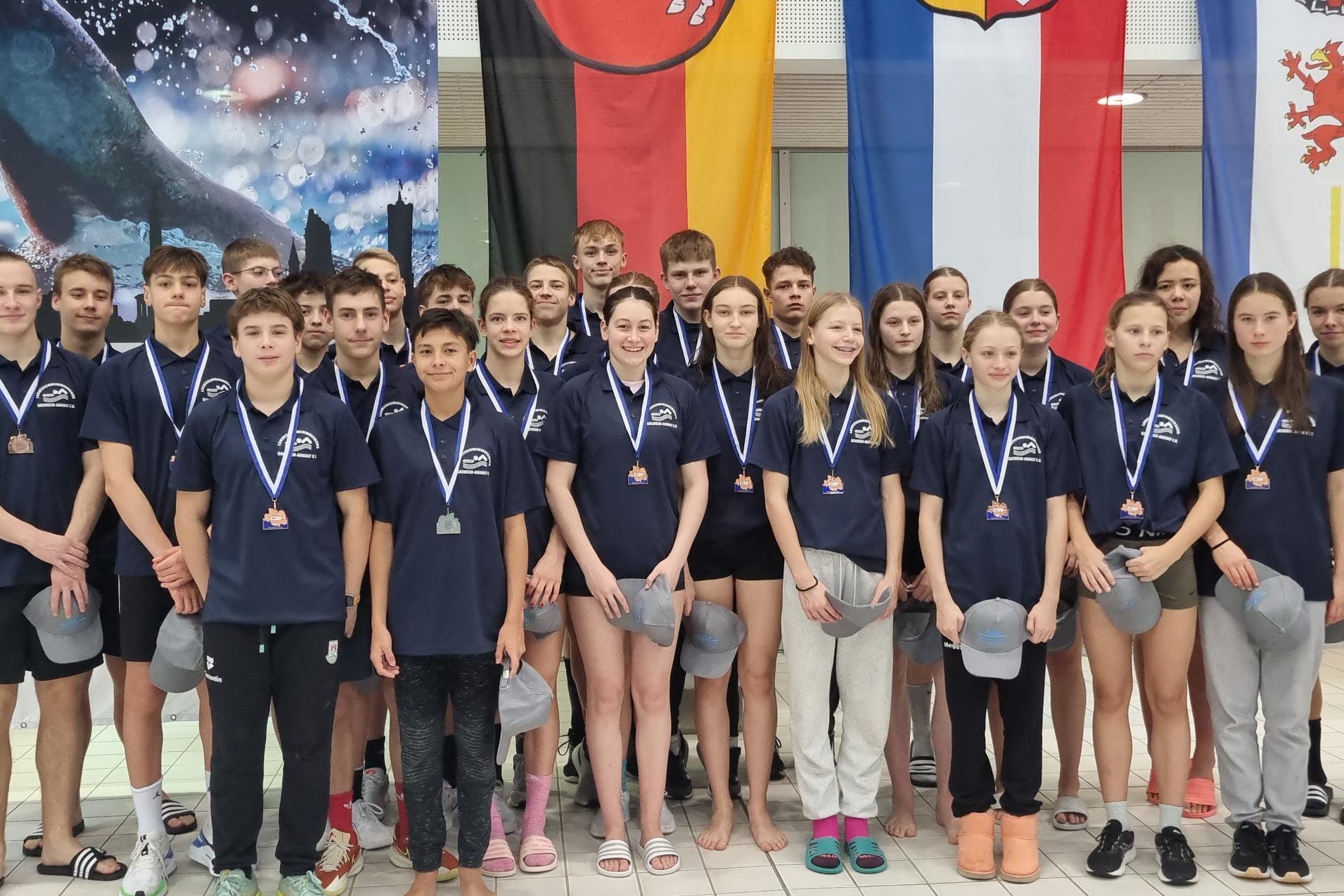 Gruppenfoto von jugendlichen Schwimmern mit Medaillen, in blauen Shirts, vor verschiedenen Flaggen in einem Schwimmbad.
