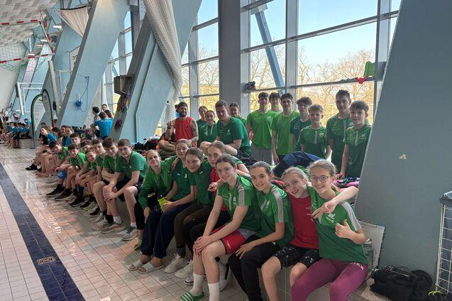 Gruppe von Schwimmern in gr&uuml;nen T-Shirts sitzt am Beckenrand eines Schwimmbads, einige halten Drinks und l&auml;cheln.