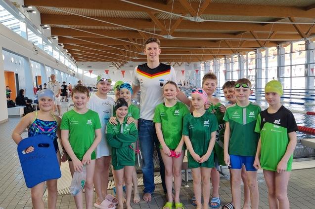Gruppe von Schwimmern in Badeanz&uuml;gen und Badekappen posiert im Hallenbad mit einem Trainer im Hintergrund.