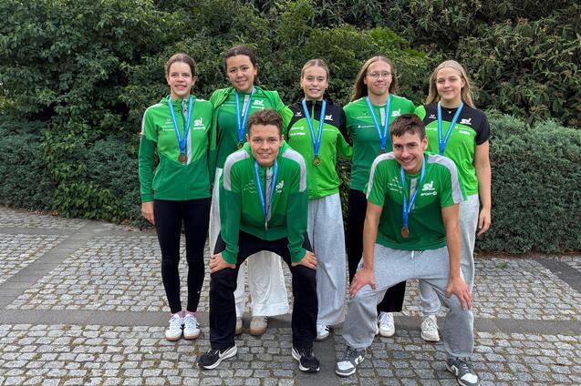 Gruppenfoto von sechs Sportlern in gr&uuml;nen Trikots mit Medaillen, posierend vor einer Hecke auf einem Pflasterweg.