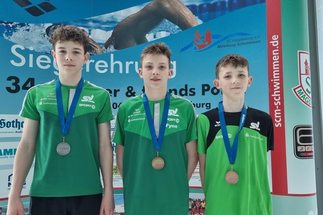 Drei junge Schwimmer mit Medaillen stehen vor einem Banner bei einer Siegerehrung im Schwimmsport.