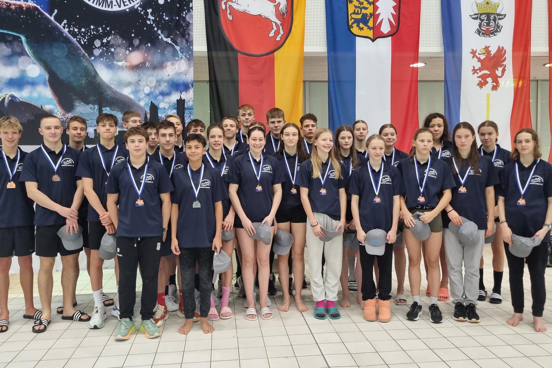 Gruppenfoto von jugendlichen Schwimmern mit Medaillen, in blauen Shirts, vor verschiedenen Flaggen in einem Schwimmbad.
