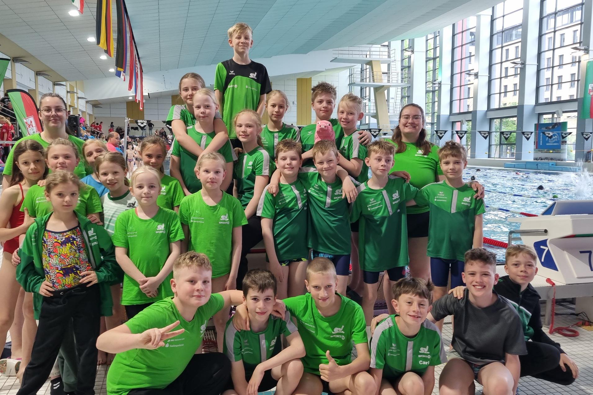 Gruppe junger Schwimmer in gr&uuml;nen T-Shirts posiert fr&ouml;hlich am Beckenrand eines Schwimmbades.