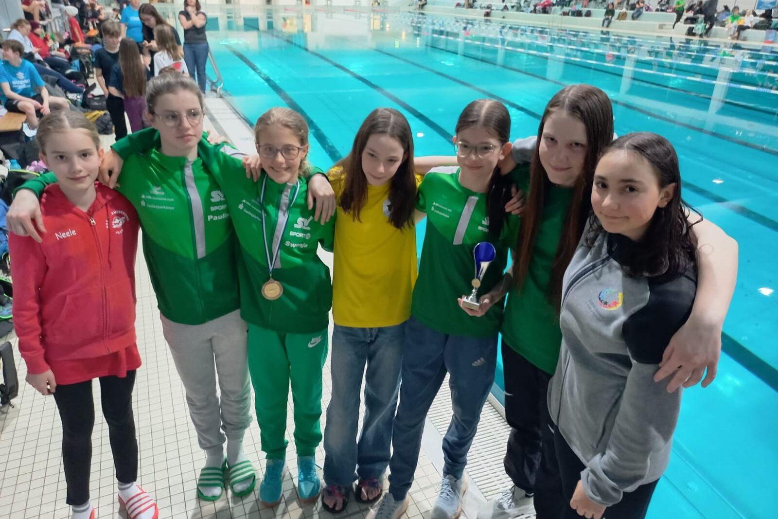 Sechs M&auml;dchen in Sportjacken stehen arm-in-arm am Schwimmbecken, eine h&auml;lt einen Pokal, andere tragen Medaillen.
