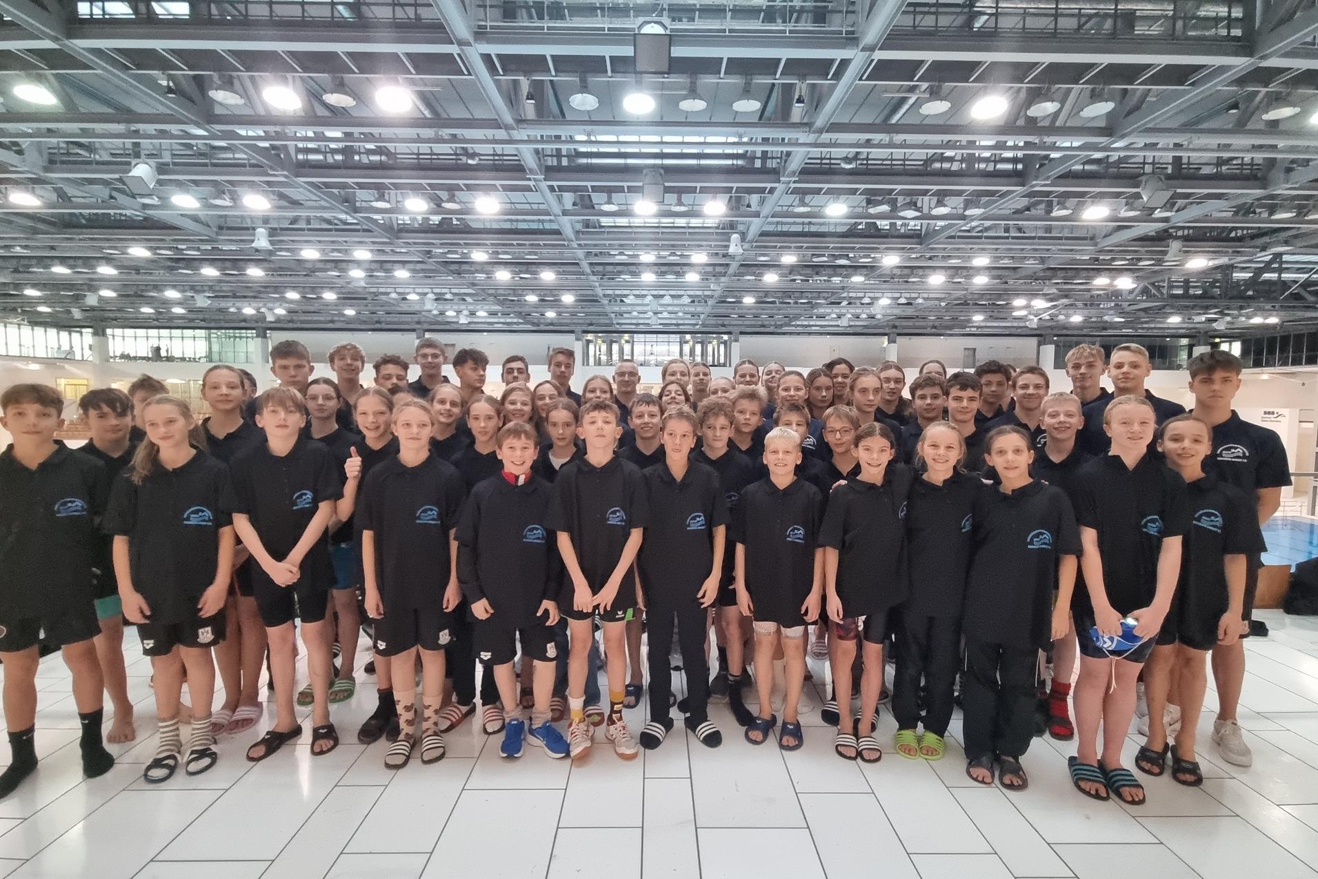 Gruppenfoto von Schwimmern in einheitlichen T-Shirts, stehend in einem modernen Schwimmbad mit Trainer im Hintergrund.