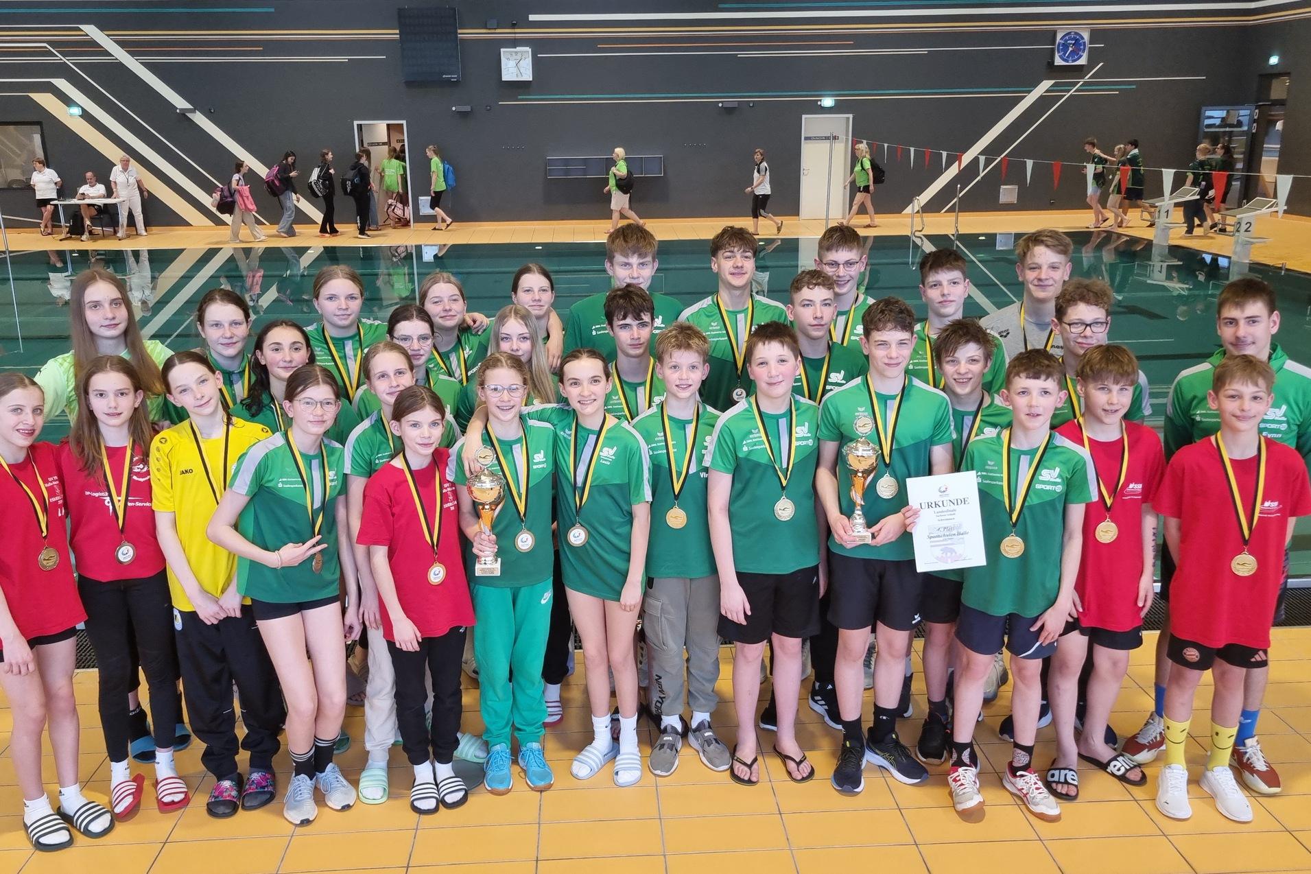 Gruppe von Schwimmern in Sportbekleidung mit Medaillen und Pokalen, posierend in einem Schwimmbad.