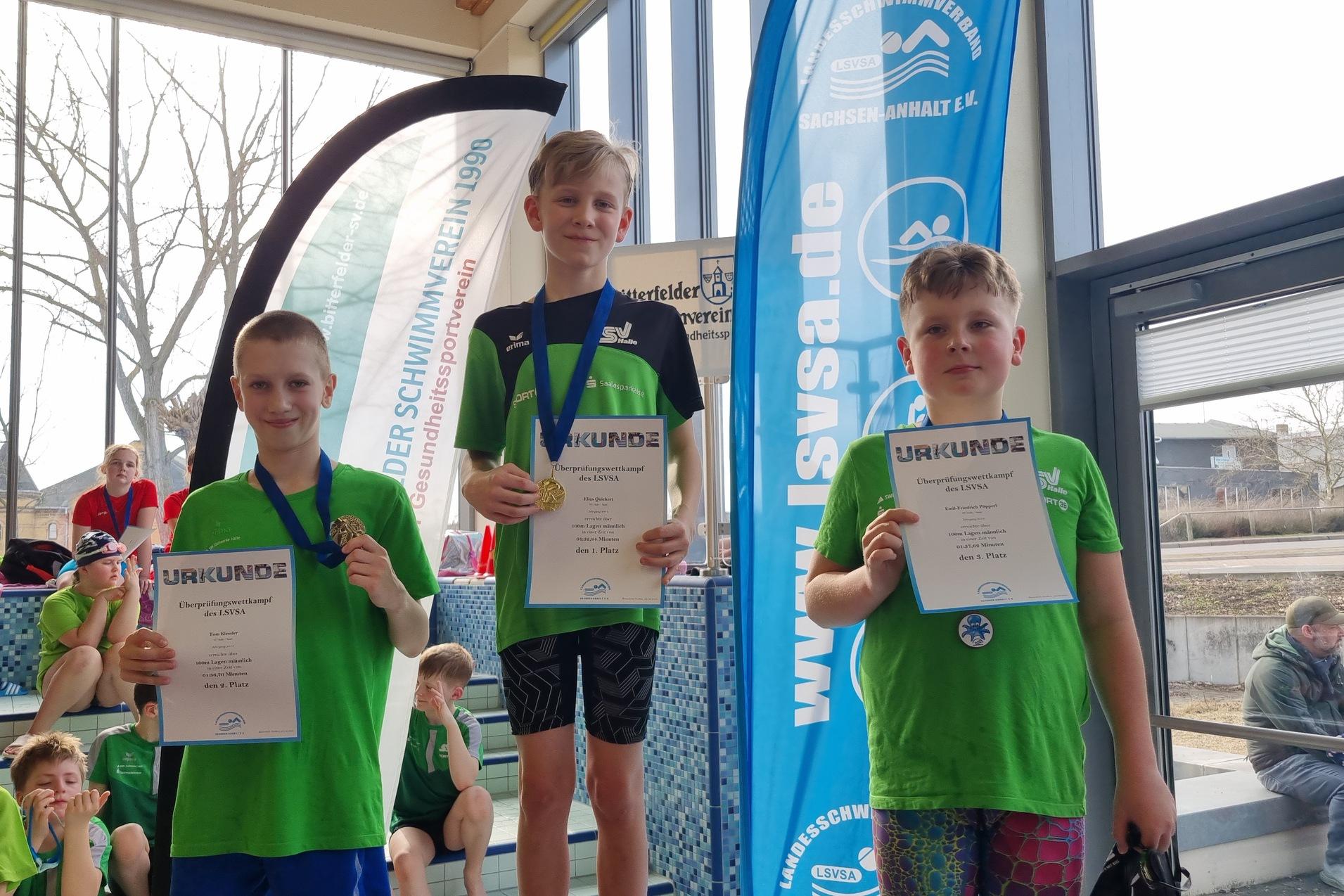 Drei Kinder in gr&uuml;nen Schwimmtrikots halten Urkunden und Medaillen bei einer Schwimmveranstaltung.