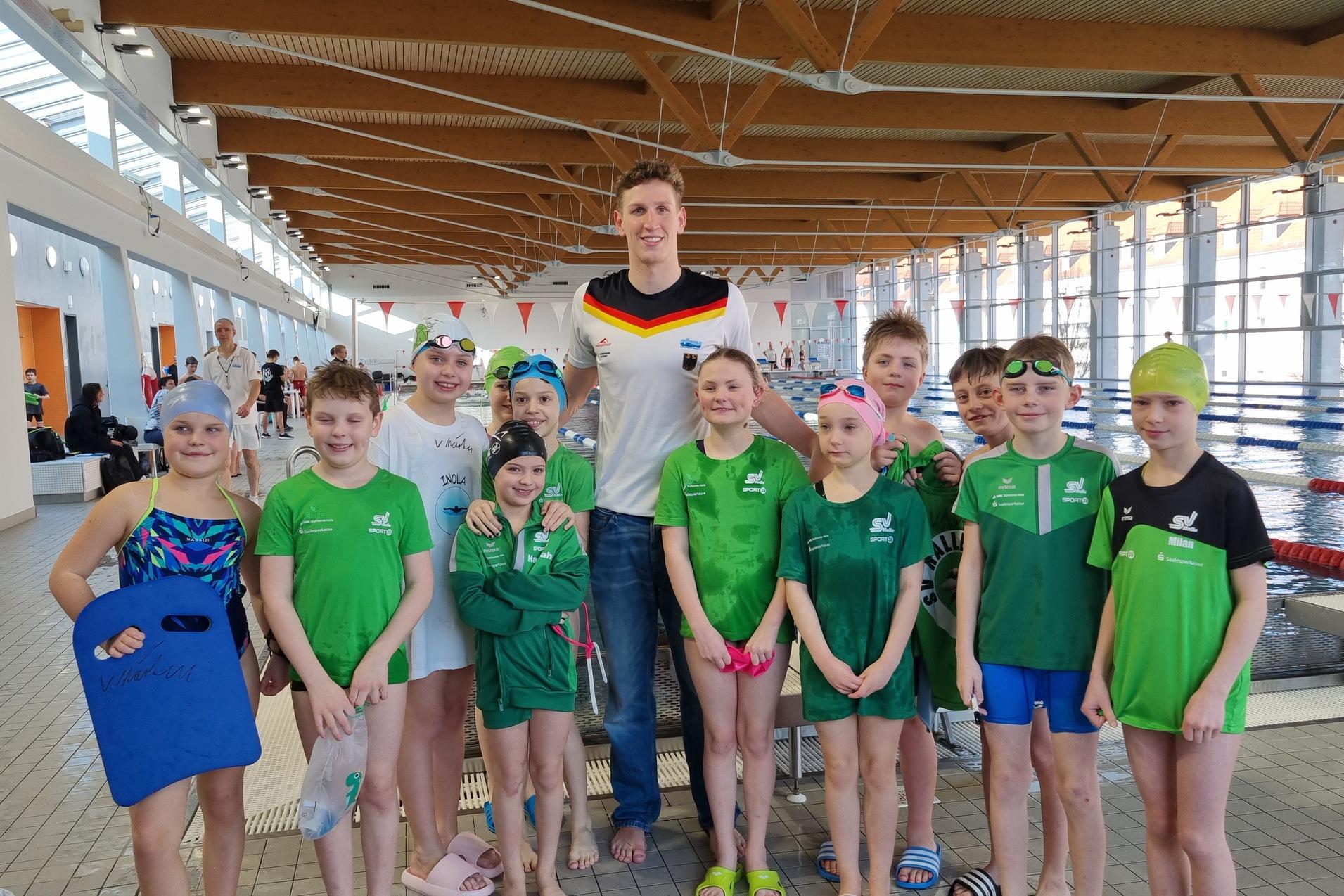 Gruppe von Schwimmern in Badeanzügen und Badekappen posiert im Hallenbad mit einem Trainer im Hintergrund.