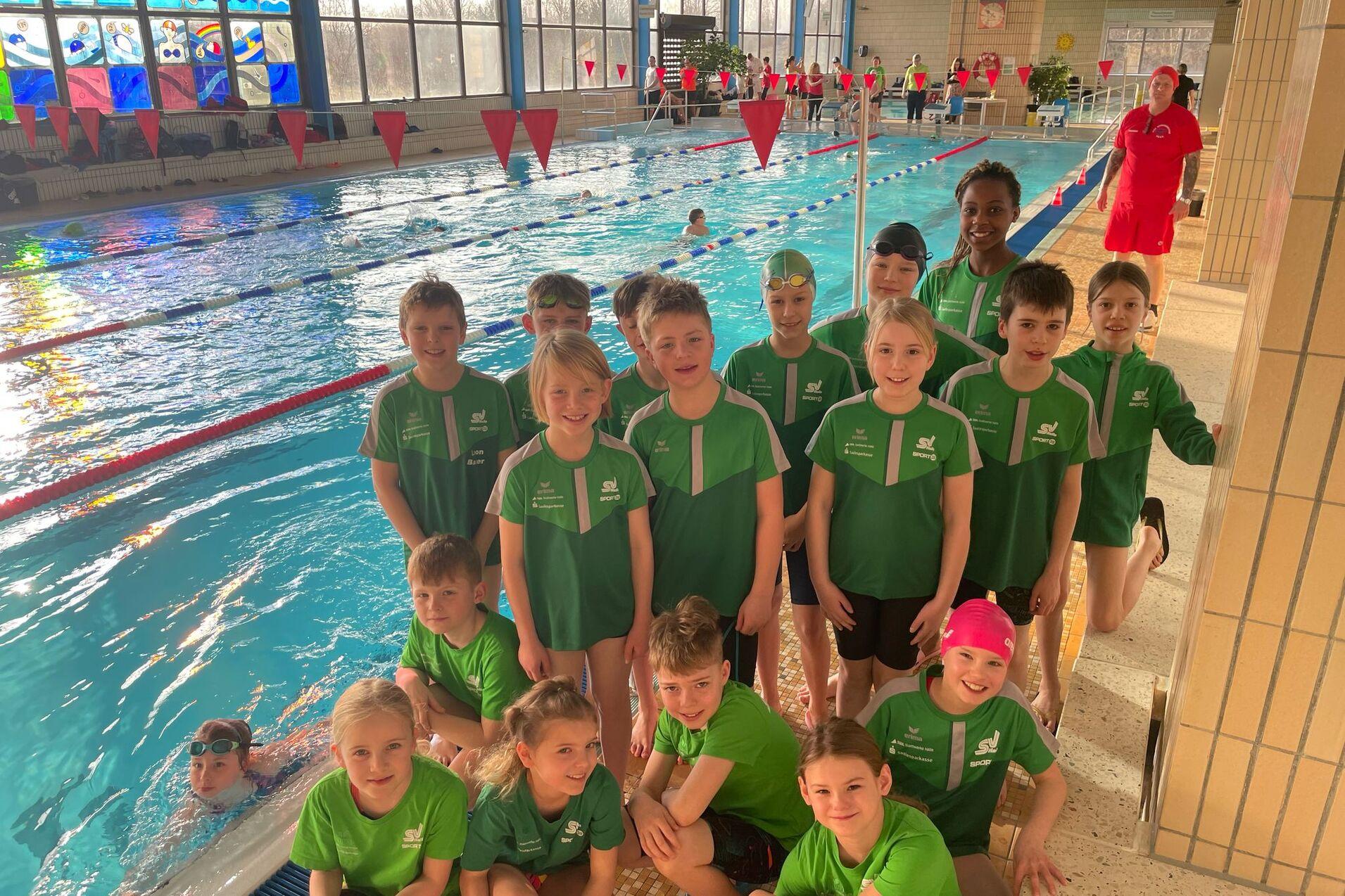 Gruppe von Kindern in grünen Schwimmanzügen steht am Beckenrand, dahinter schwimmen weitere Kinder im Pool.