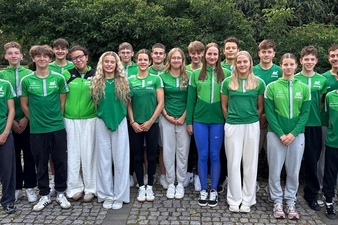 Gruppenbild von 16 jungen Sportlern in gr&uuml;nen Trikots, stehend vor einer gr&uuml;nen Hecke auf einem gepflasterten Weg.