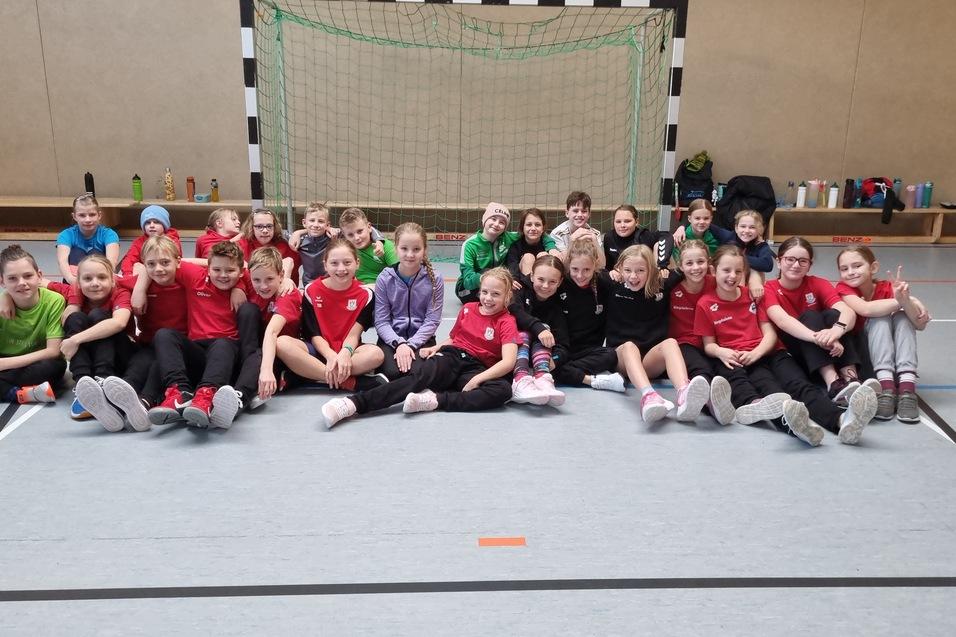 Gruppenfoto von Kindern in Sportkleidung, die auf dem Boden einer Halle vor einem Handballtor sitzen.