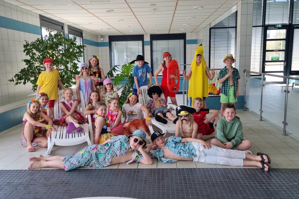 Gruppenfoto von Kindern in bunten Kost&uuml;men und Sonnenaccessoires, posierend im Schwimmbad mit Pflanzen im Hintergrund.