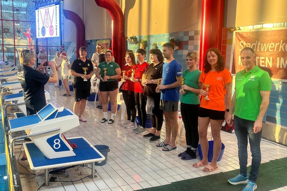 Medailleverleihung mit Teilnehmern in Sportkleidung, die Pokale in der Hand halten, vor einer Schwimmhalle.