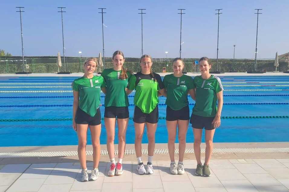 F&uuml;nf Schwimmerinnen in gr&uuml;nen T-Shirts stehen am Rand eines Schwimmbeckens unter blauem Himmel.