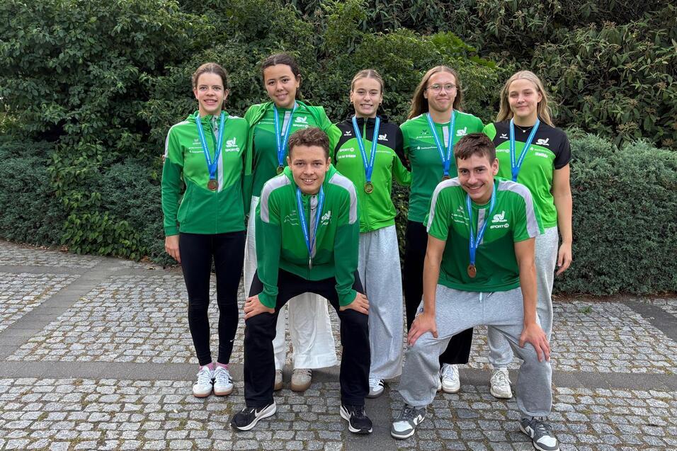 Gruppenfoto von sechs Sportlern in grünen Trikots mit Medaillen, posierend vor einer Hecke auf einem Pflasterweg.