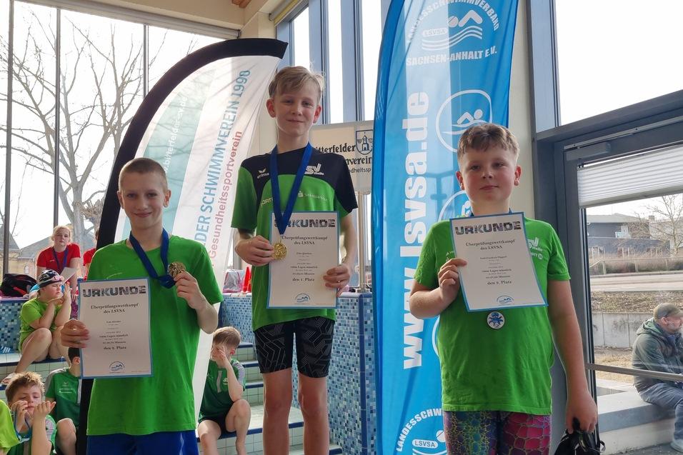 Drei Kinder in gr&uuml;nen Schwimmtrikots halten Urkunden und Medaillen bei einer Schwimmveranstaltung.