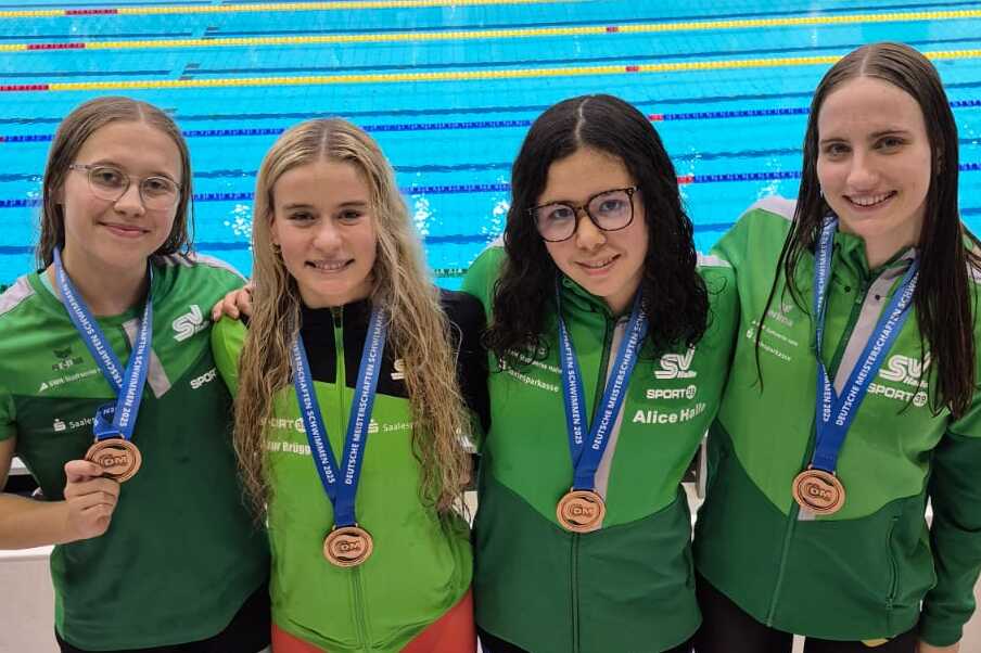 Vier Schwimmerinnen in gr&uuml;nen Trikots mit Medaillen posieren vor einem Wettkampfbecken.