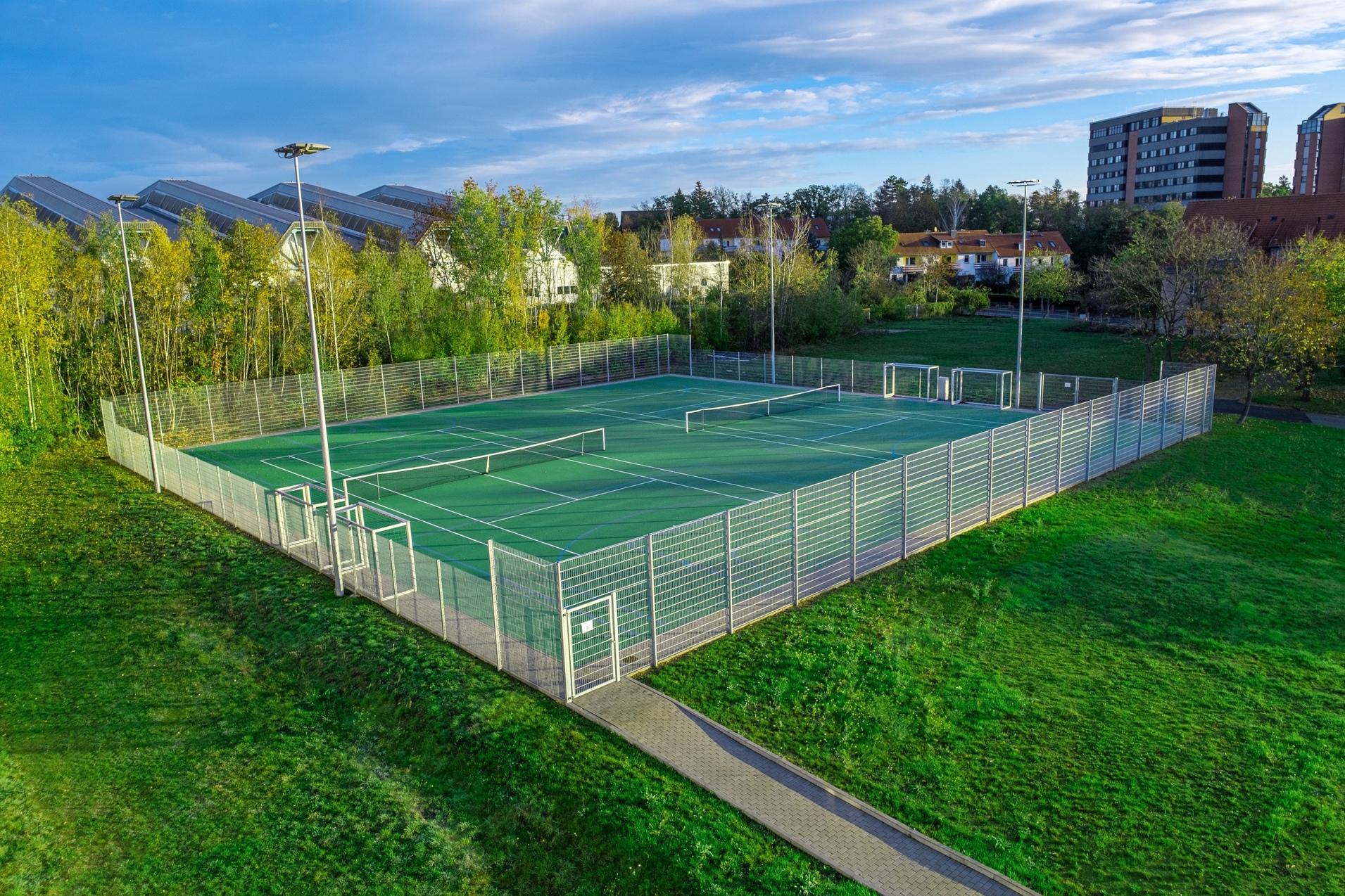 Luftaufnahme eines Tennisplatzes mit Netz, umgeben von Zaun und Grünflächen, im Hintergrund Wohngebäude.