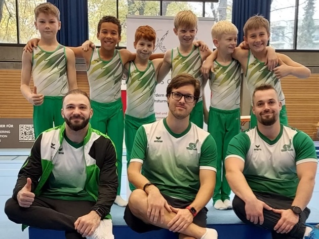 Gruppe von sieben männlichen Turnern in Sportkleidung posieren fröhlich auf einer Turnmatte bei einem Wettkampf.