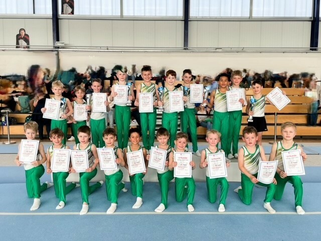 Gruppe von jungen Sportlern in grünen Trikots, die stolz ihre Urkunden in einer Turnhalle präsentieren.
