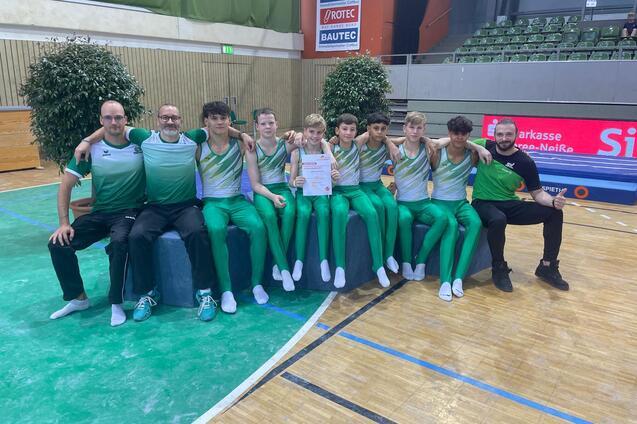 Gruppenfoto von Gymnastikern in gr&uuml;nen Anz&uuml;gen, umgeben von Trainern, mit Urkunde in der Mitte.