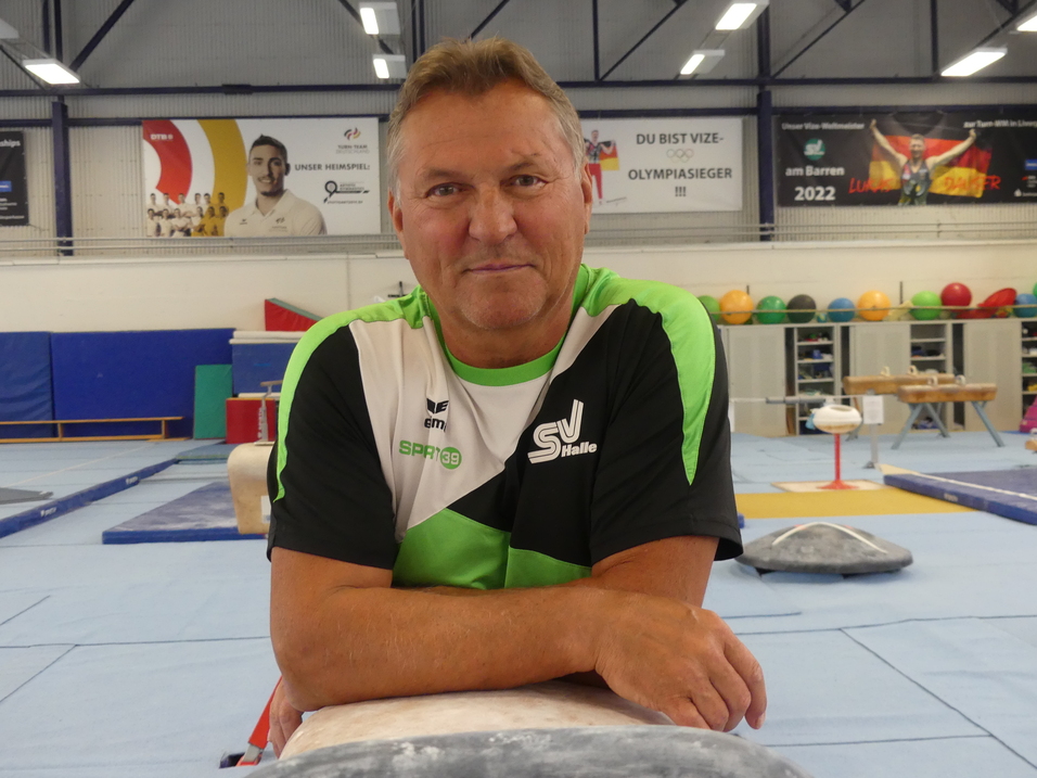 Mann in Sportkleidung lehnt auf einem Turngerät in einer Turnhalle mit Barren und Gymnastikgeräten im Hintergrund.