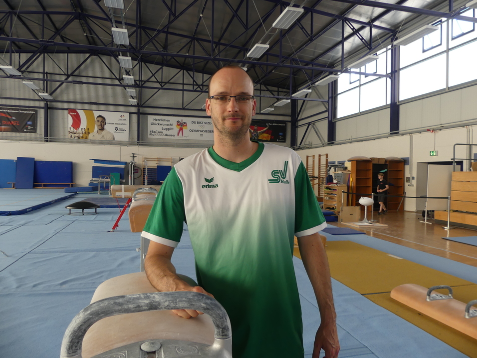 Mann in Sportkleidung steht an einem Turngerät in einer Sporthalle mit blauen Matten und weiteren Geräten im Hintergrund.