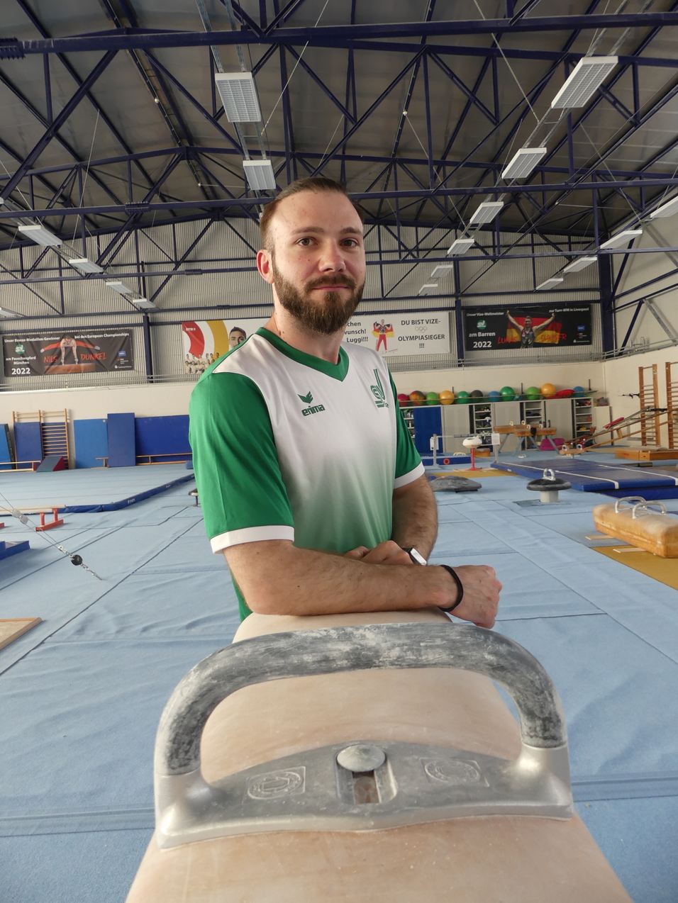 Mann mit Bart steht an einem Barren in einer Sporthalle mit Turngeräten im Hintergrund.