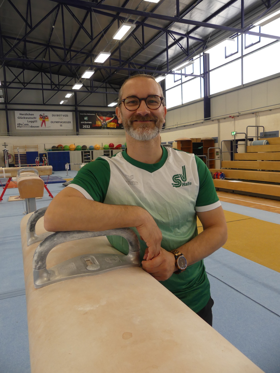 Lächelnder Trainer in Sportkleidung lehnt an einem Turnpferd in einer Sporthalle mit diferentes Geräten im Hintergrund.