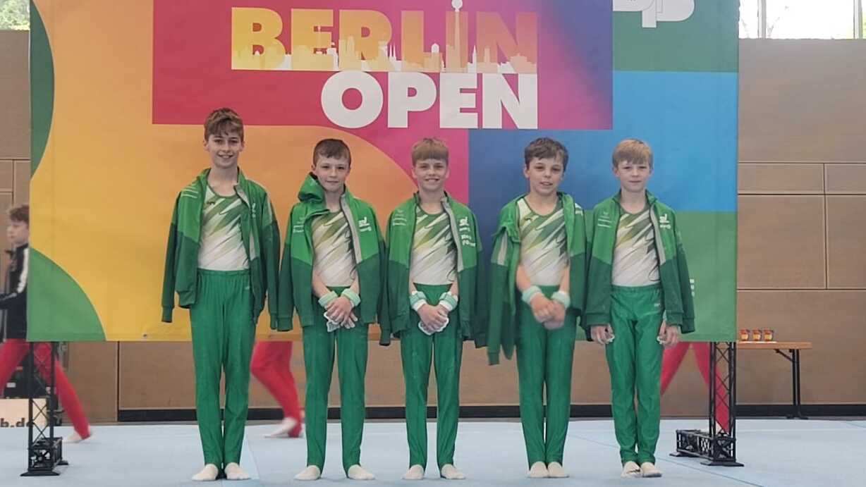Fünf Kinder in grünen Sportanzügen stehen vor einem bunten Banner der Berlin Open in einer Sporthalle.