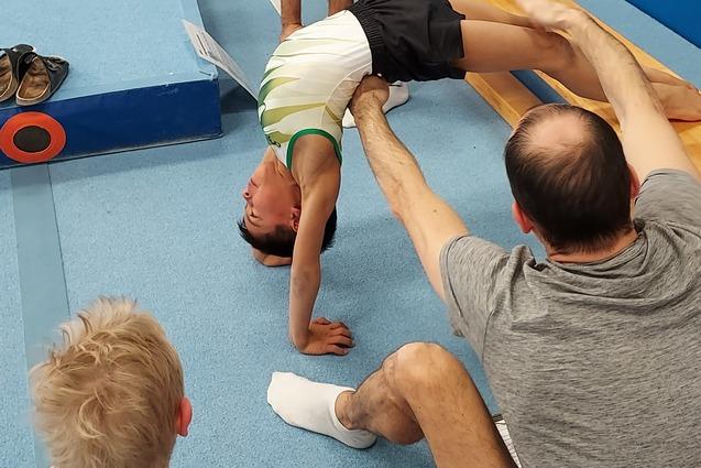 Ein Junge macht eine Br&uuml;cke, w&auml;hrend Trainer und Zuschauer im Gymnastikraum zuschauen. Matte und Turnger&auml;te im Hintergrund.