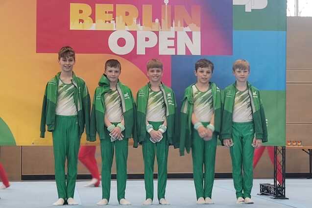 F&uuml;nf Kinder in gr&uuml;nen Sportanz&uuml;gen stehen vor einem bunten Banner der Berlin Open in einer Sporthalle.