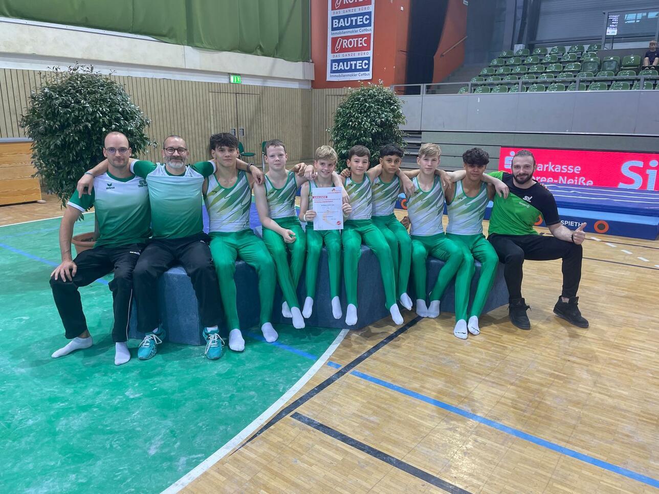 Gruppenfoto von Gymnastikern in grünen Anzügen, umgeben von Trainern, mit Urkunde in der Mitte.