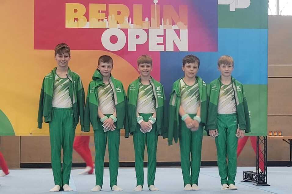 Fünf Kinder in grünen Sportanzügen stehen vor einem bunten Banner der Berlin Open in einer Sporthalle.