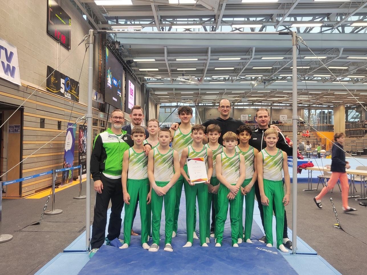 Gruppe von Jungen in Sportkleidung und Trainern, posierend mit einer Urkunde an einem Gymnastikgerüst.