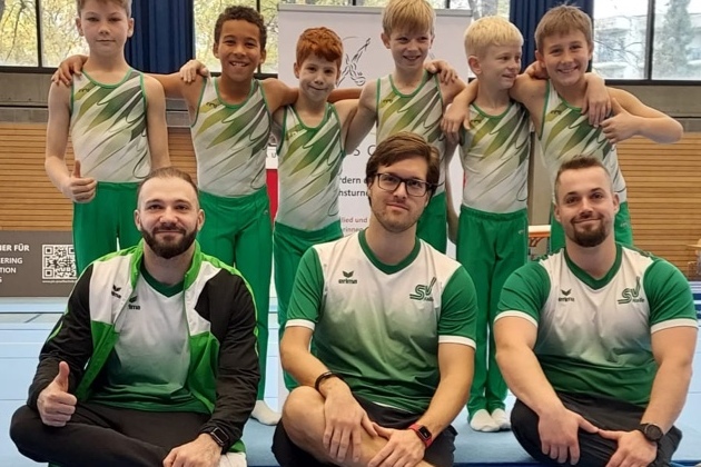 Gruppe von sieben m&auml;nnlichen Turnern in Sportkleidung posieren fr&ouml;hlich auf einer Turnmatte bei einem Wettkampf.