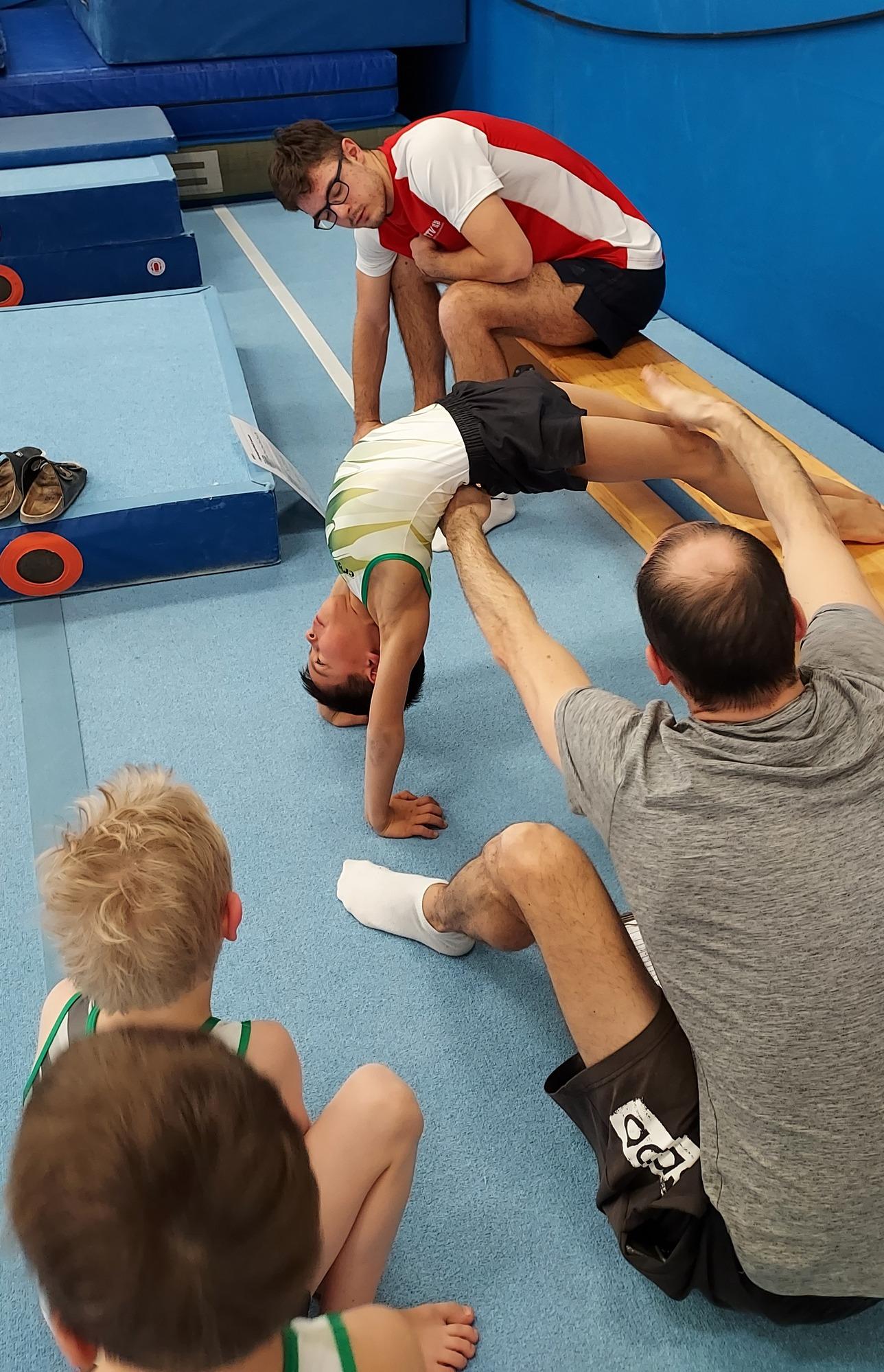 Ein Junge macht eine Brücke, während Trainer und Zuschauer im Gymnastikraum zuschauen. Matte und Turngeräte im Hintergrund.