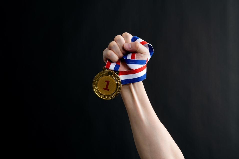 Eine Hand hält eine goldene Medaille mit der Nummer 1 und einem rot-blauen Band vor dunklem Hintergrund.