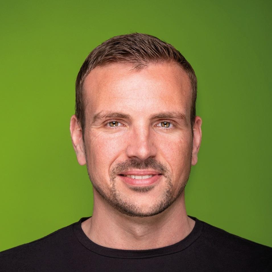 Porträt eines Mannes mit kurzem Haar und Bart, der ein schwarzes T-Shirt trägt, vor grünem Hintergrund.