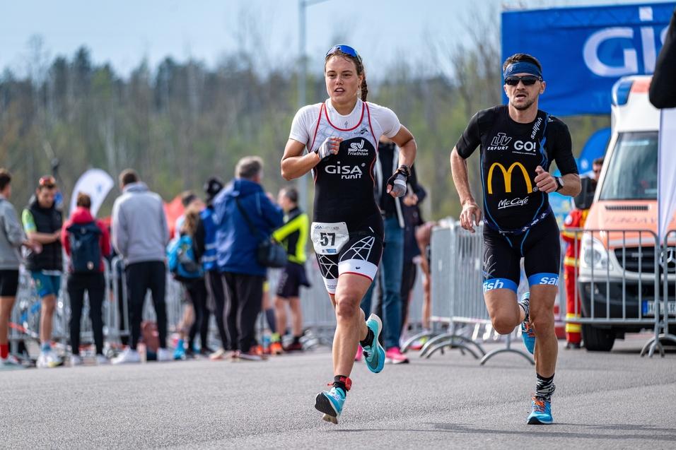 Zwei Athleten, ein Mann und eine Frau, laufen bei einem Triathlon durch eine Zuschauergruppe. Im Hintergrund sind Flaggen.