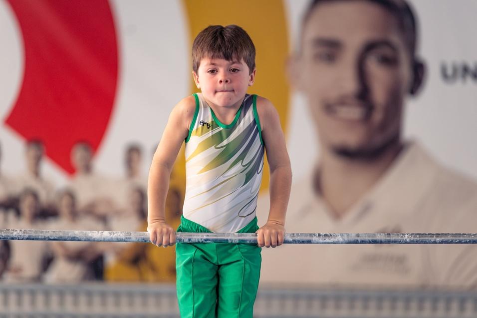 Ein Junge in einem Gymnastikanzug übt an einem Reck in einer Sporteinrichtung mit buntem Hintergrund.