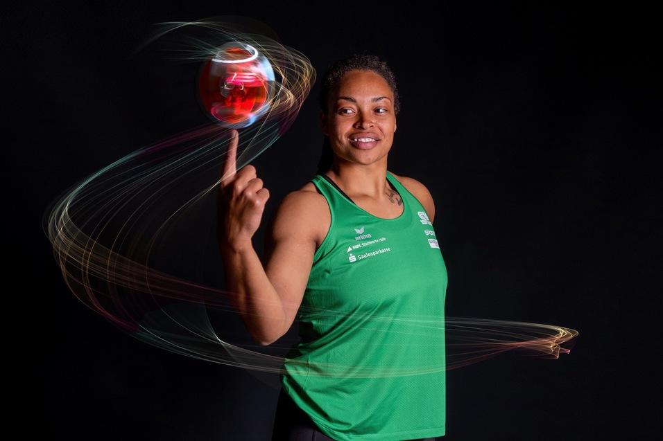 Sportlerin in gr&uuml;nem Trikot balanciert einen roten Ball auf dem Zeigefinger, mit dynamischen Lichtspuren im Hintergrund.
