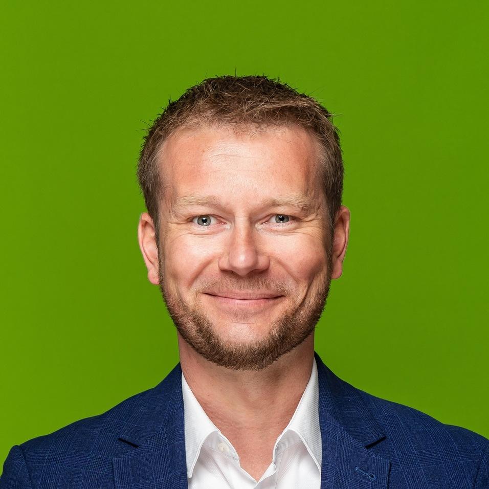 Porträt eines Mannes mit kurzem Haar und Bart, der in einem blauen Blazer und weißem Hemd vor einem grünen Hintergrund lächelt.