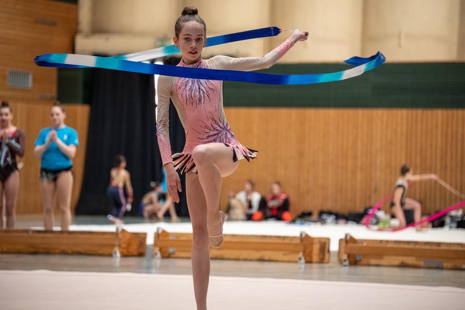 Mädchen in einem bunten Gymnastikanzug führt einen Tanz mit einem blauen Band auf einer Sportfläche aus.