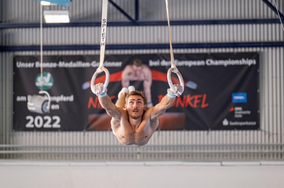 Mann in der Luft, der an Ringen hängt, mit einem Banner im Hintergrund für die Europameisterschaften.