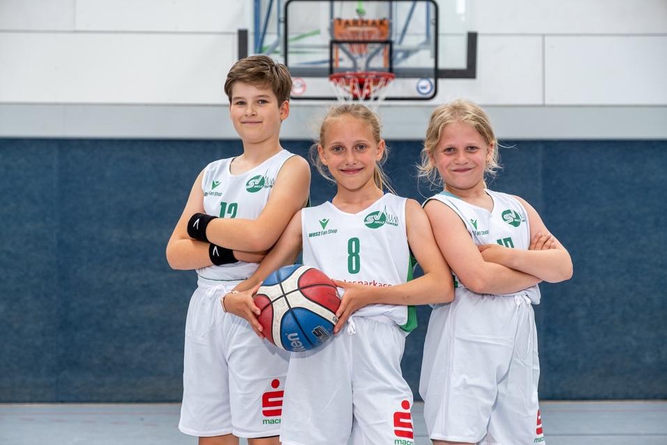 Drei Kinder in Basketball-Trikots stehen lächelnd vor einem Basketballkorb und halten einen Ball.