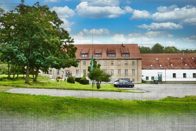 Historisches Gebäude mit roten Dächern, angrenzender Grünfläche und Fahrzeugen auf einem Weg. Klarer Himmel mit Wolken.