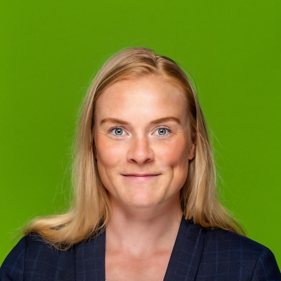 Porträt einer Frau mit blonden Haaren, die ein dunkelblaues, kariertes Sakko und ein weißes Shirt trägt, vor grünem Hintergrund.